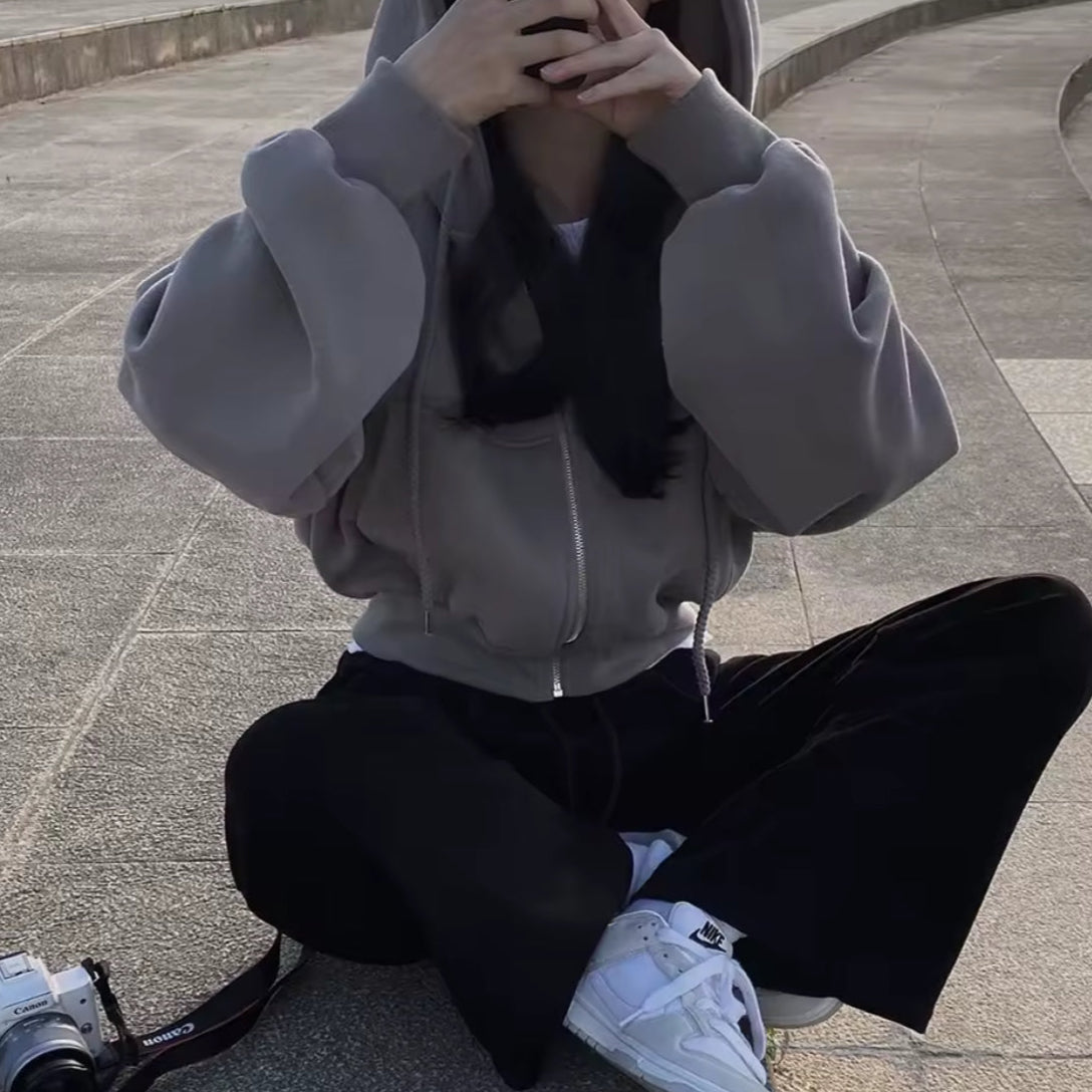 Person sitting on the ground wearing a gray hoodie and black pants with white sneakers, holding a camera.