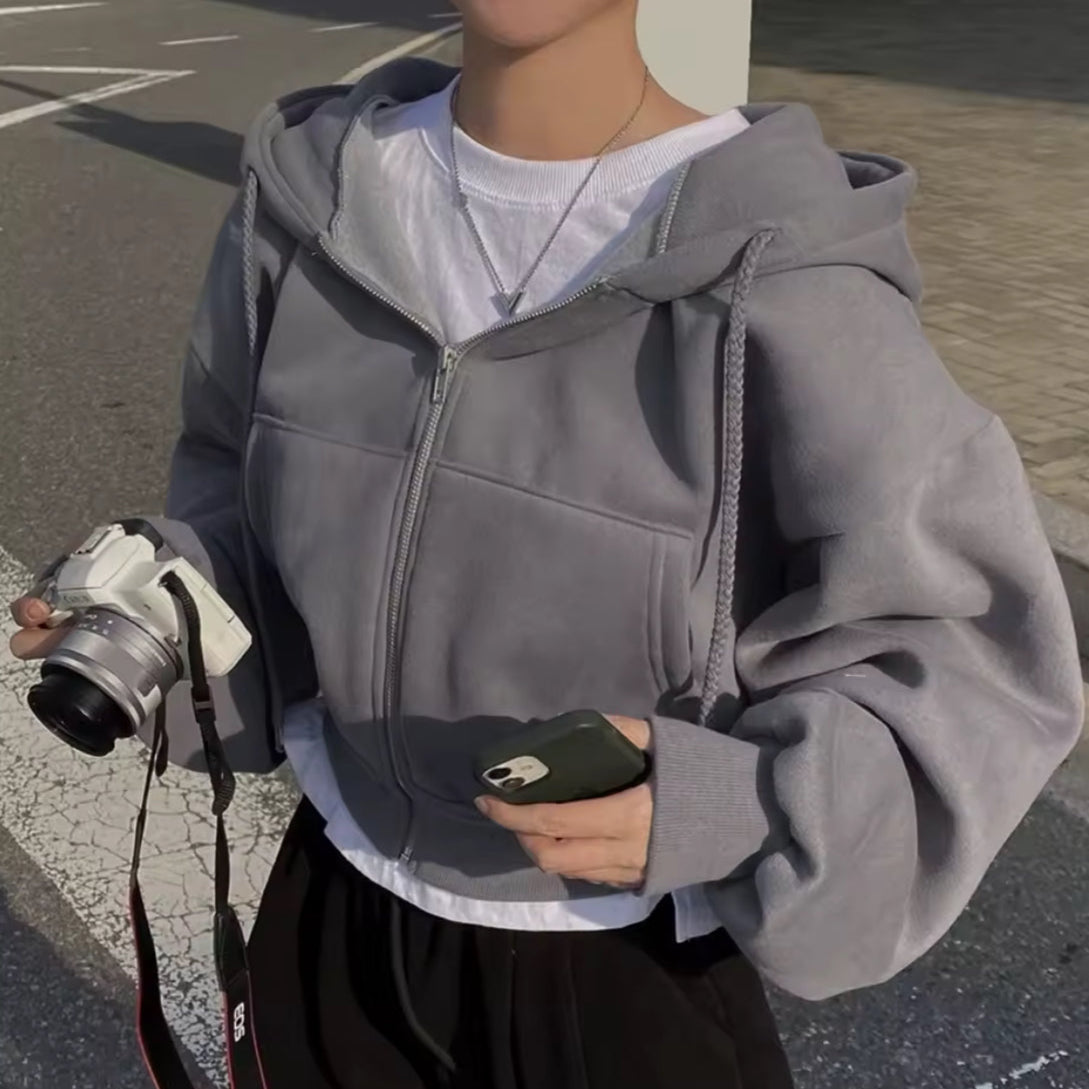 Person wearing a gray hoodie holding a camera and phone on a street.